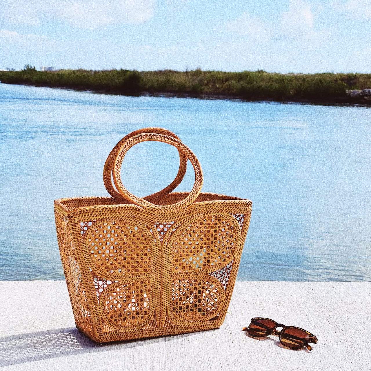The Butterfly Rattan Handbag handmade by Ganapati Crafts Co. in Bali is sitting on a rock with sunglasses lying near to it looking stylish