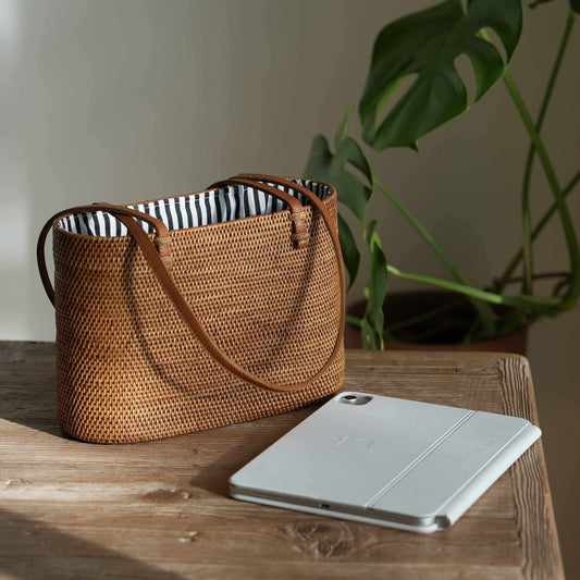 A detailed, eye-level shot of a rectangular, handwoven rattan tote bag resting on a rustic wooden table. The bag, golden-brown in the natural sunlight, features thin, tan leather shoulder straps looped over the sides. The open top reveals a crisp, black-and-white vertically striped fabric lining. In the foreground to the right, a silver tablet with a folded white magnetic cover lies on the table. The scene is illuminated by soft light from the left, casting a gentle shadow on the plain wall behind.