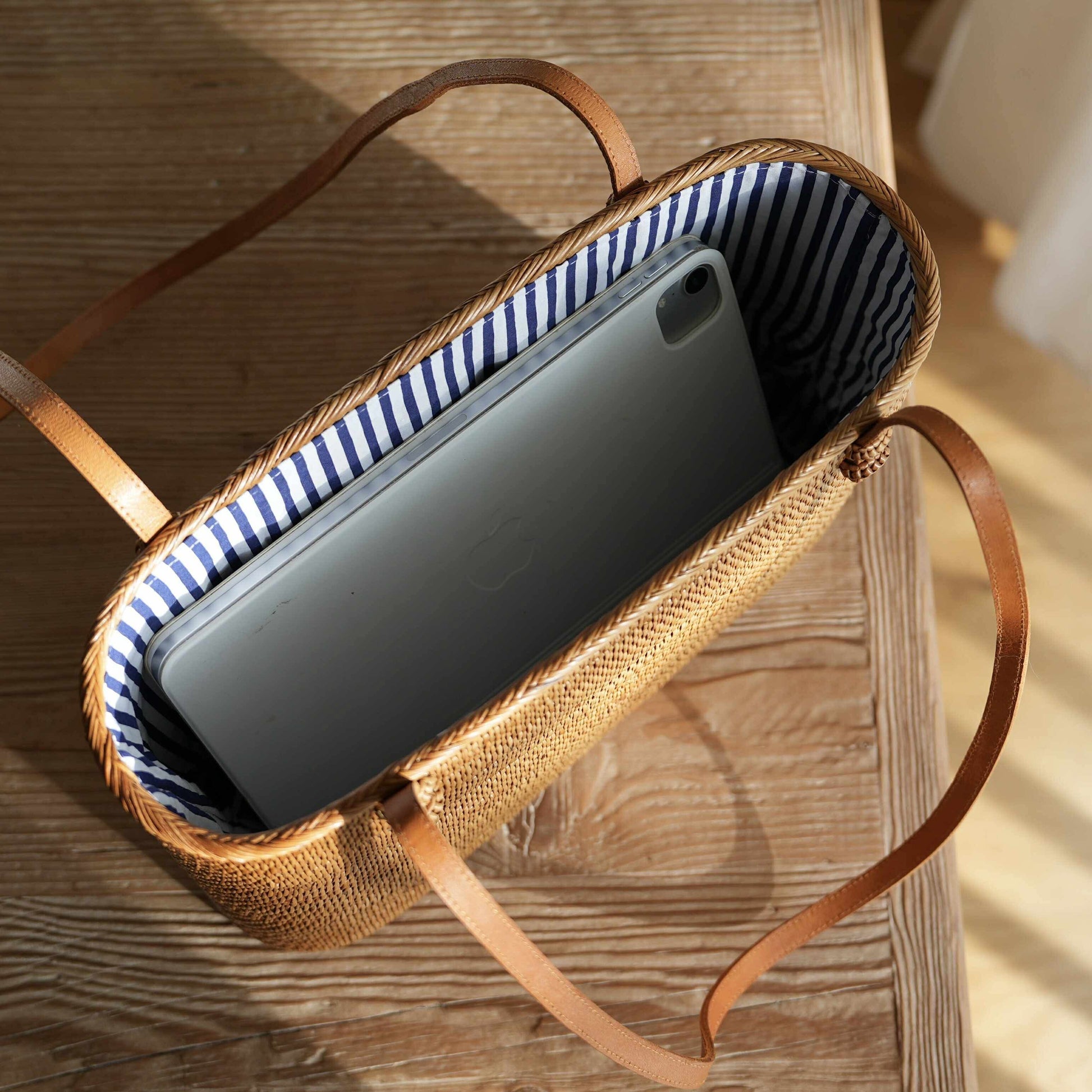 A high-angle, close-up photograph shows a handwoven, rectangular rattan tote bag resting on a rustic wooden table. The bag is filled with a silver tablet, demonstrating its size and capacity. The interior of the golden-brown bag is lined with a blue-and-white striped fabric, and its thin, tan leather shoulder straps rest on the table. Strong natural light from the left highlights the intricate weave of the rattan and creates a bright glare on the tablet's screen.