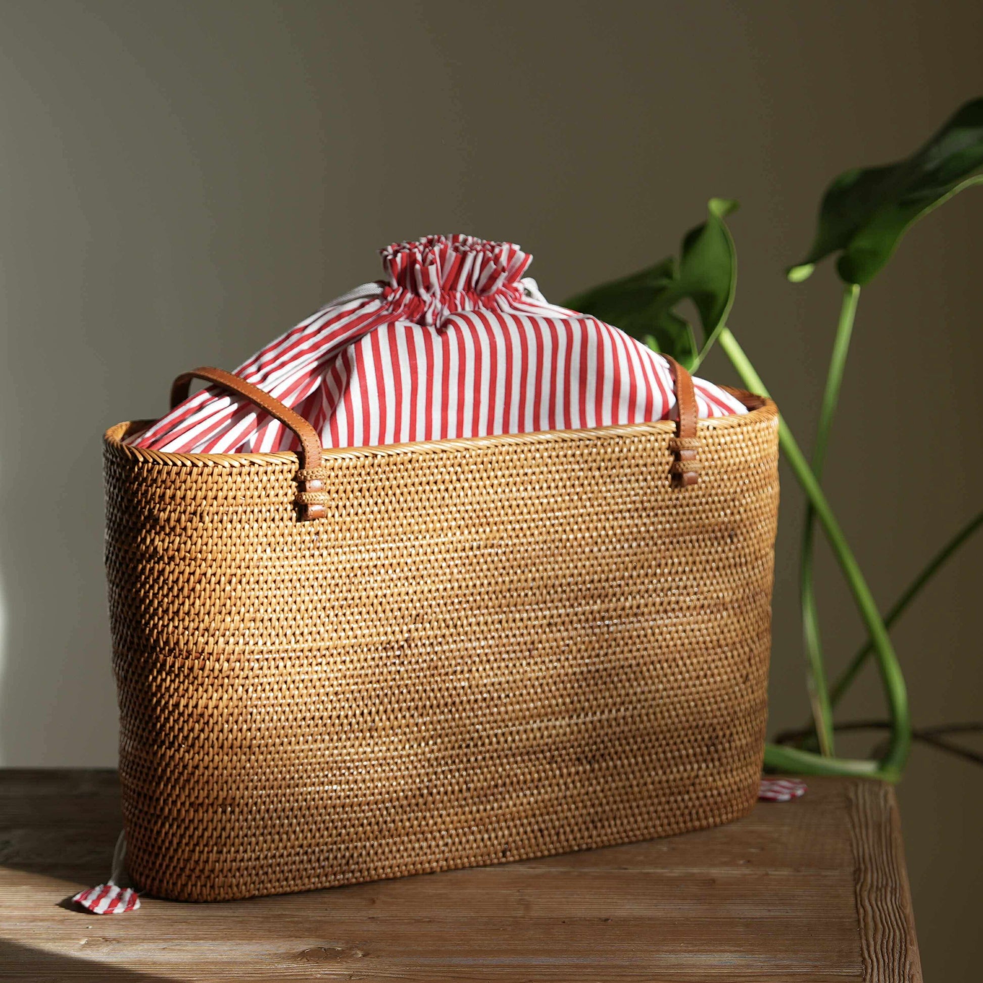 A handwoven rattan tote bag sits in a pool of natural sunlight on a rustic wooden table. The bag features elegant tan leather handles and a vibrant red-and-white striped fabric lining, which is cinched closed with a drawstring at the top. In the background, the soft green leaves of a monstera plant add a touch of nature, complementing the bag's organic materials and exquisite craftsmanship.