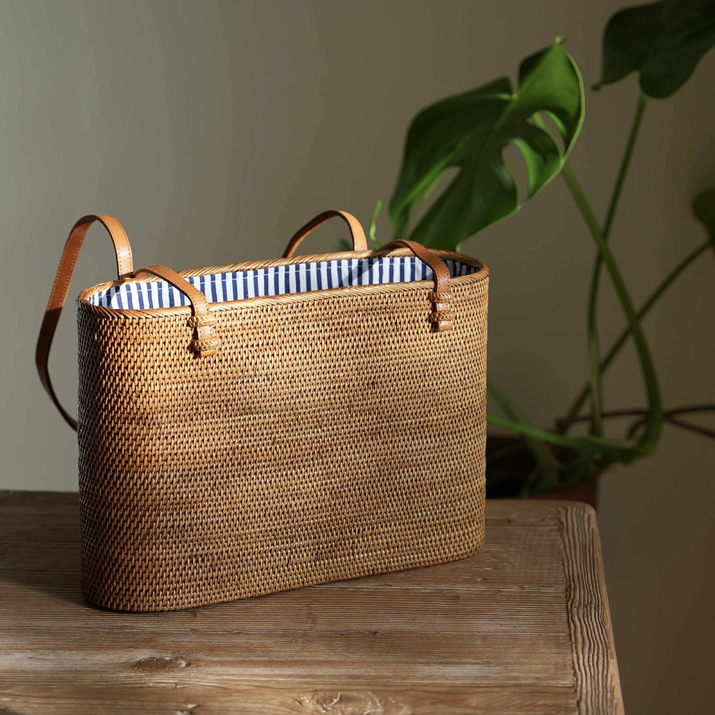 This beautifully handcrafted Bali Rattan Shoulder Bag, made from natural woven Ata grass, rests on a rustic wooden table in a sunlit room. Its classic tote design is elevated by rich, brown leather handles and a charming blue-and-white striped interior lining. Ethically handmade in Bali, this eco-friendly bag combines timeless style with sustainable craftsmanship, making it a perfect accessory for any occasion. A monstera plant in the background enhances its natural, artisanal appeal.