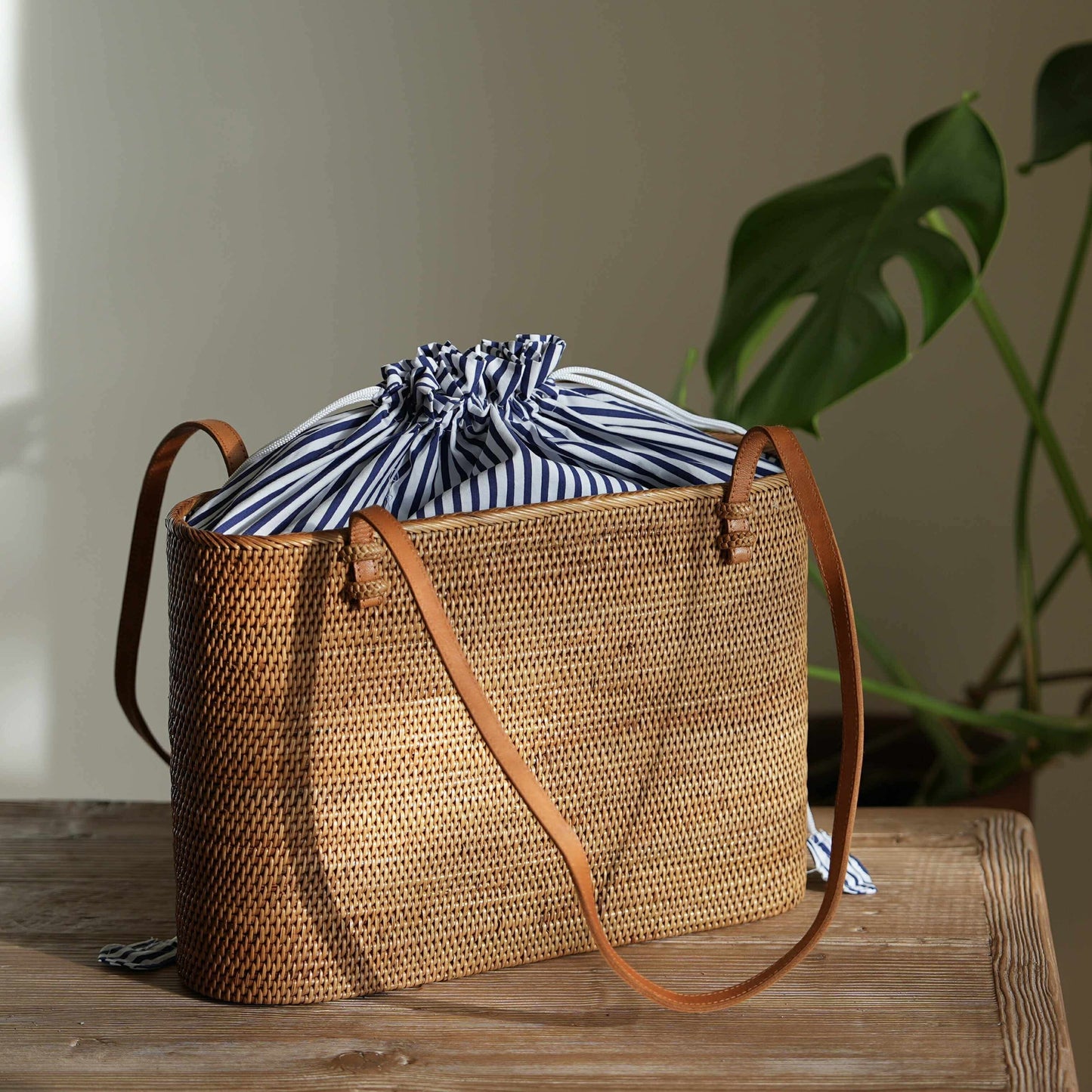 A rectangular, handwoven rattan tote bag sits on a rustic wooden table in a warmly lit room. The bag has thin, brown leather shoulder straps attached to the top rim. Peeking out from the top is a blue and white striped fabric lining, which is gathered and tied shut with a white drawstring. Sunlight from the left highlights the texture of the weave and casts a soft shadow behind it. In the blurred background, the large, dark green leaves of a monstera plant are visible.