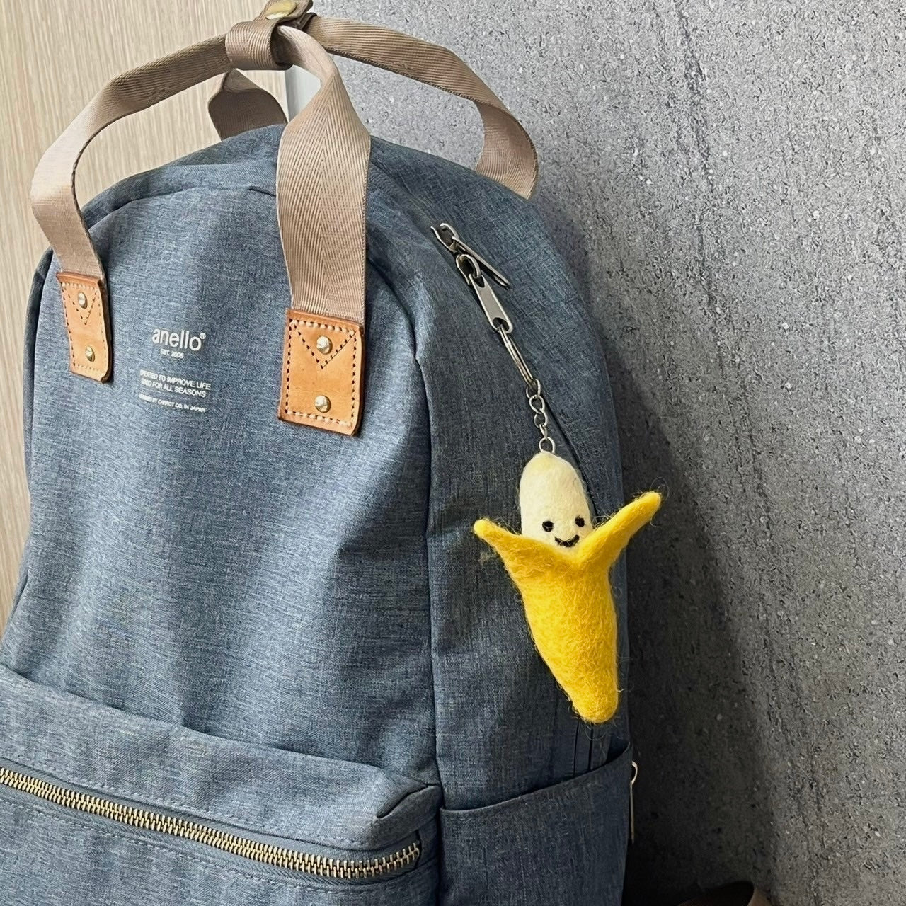A cheerful handmade felt smiling banana keychain is attached to a textured grey blue backpack. The whimsical yellow banana, featuring a white top and a smiling face, adds a playful touch. The grey backpack, with its brown straps and leather accents, is casually hung, ready for everyday adventures.